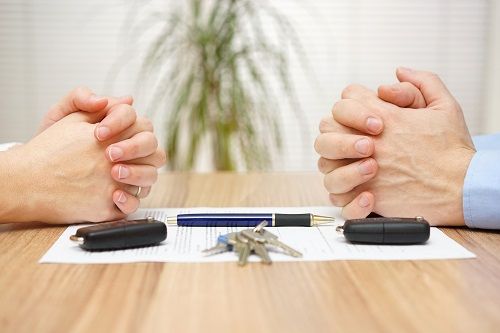 A pair of folded hands across from each other on a table with a piece of paper, pen, two key fobs and a set of keys.